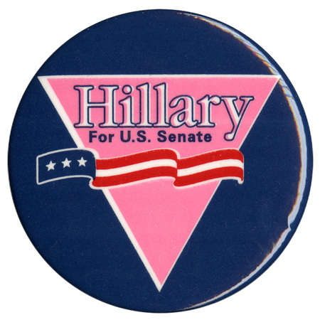 Round, blue button with an inverted pink triangle and a ribbon with red, white, and blue stars and stripes. White and blue text reads "Hillary For U.S. Senate"