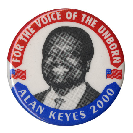 Round, white button with a black and white photograph of Alan Keyes flanked by color illustrations of the US flag. In the red and blue border of the button is white text reading "FOR THE VOICE OF THE UNBORN ALAN KEYES 2000"