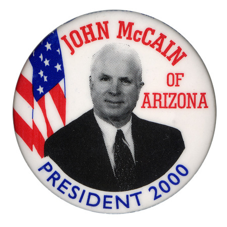 Round, white button with a black and white photograph of John McCain and a color illustration of the US flag. Red and blue text reads "JOHN McCAIN OF ARIZONA PRESIDENT 2000"
