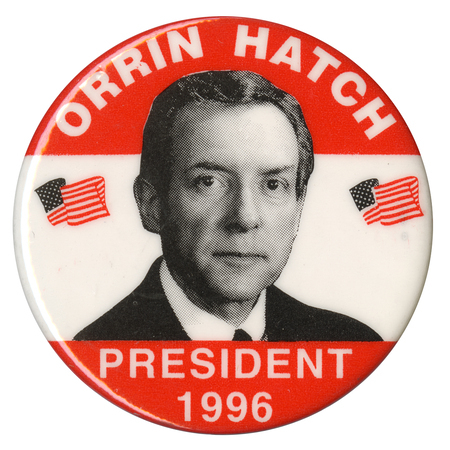 Round button with bands of white and orange. A black and white photograph of Orrin Hatch is in the center, between two drawings of the US flag. White text reads "ORRIN HATCH PRESIDENT 1996"