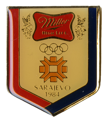 Shield-shaped gold pin with a red and blue border. In the middle is a red patch with gold text reading "Miller High Life". Below it are the five interlocking gold rings symbol of the Olympic Games, the orange blocky logo of the Sarajevo games, and gold text reading "SARAJEVO 1984"