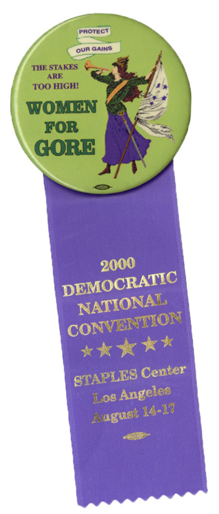 Round, green button with a color illustration of a woman in a blue skirt, green shirt, and yellow sash holding a white flag with blue stars and blowing a golden horn. Black and green text reads "PROTECT OUR GAINS THE STAKES ARE TOO HIGH! WOMEN FOR GORE". Attached to the bottom of the button is a purple ribbon with gold text reading "2000 DEMOCRATIC NATIONAL CONVENTION STAPLES Center Los Angeles August 14-17"
