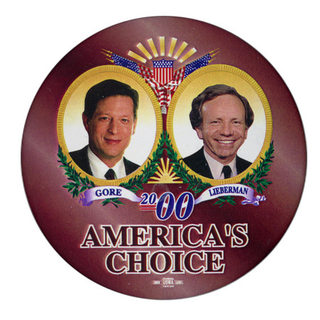Round, brown button with color photographs of Al Gore and Joe Lieberman in elaborate frames with US flags, greenery, a shield, and gold rays. Blue and black text reads "GORE LIEBERMAN 2000 AMERICA'S CHOICE"