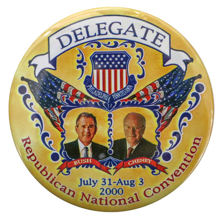 Round, yellow button with color photographs of George W. Bush and Dick Cheney in an elaborate frame with US flags, a shield, and blue curlicues. Under the photographs is white text reading "BUSH CHENEY". Under the shield is white text reading "PHILADELPHIA, PENNSYLVANIA." Around the edge of the button is black and red text reading "DELEGATE Republican National Convention July 31-Aug 3 2000"