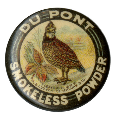 Round button with a black border surrounding a color illustration of a quail among leaves and wood. White text around the edge reads "DU PONT SMOKELESS POWDER"