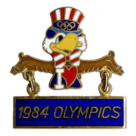 Enameled button in the shape of Sam the Olympic Eagle, a bald eagle wearing a red, white, and blue top hat and bowtie, holding a rectangular blue banner. White, red, and gold text reads "I [heart shape] 1984 OLYMPICS"