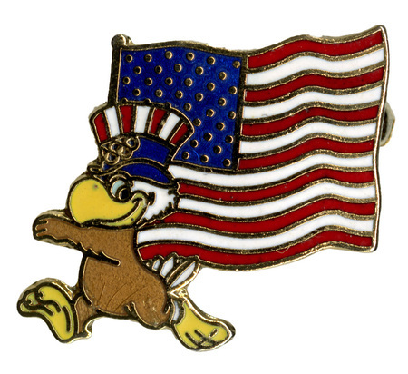 Enameled button in the shape of Sam the Olympic Eagle, a bald eagle wearing a red, white, and blue top hat and bowtie, holding US flag.