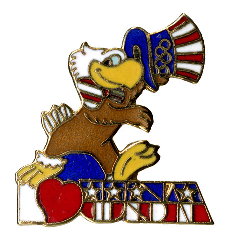 Enameled button in the shape of Sam the Olympic Eagle, a bald eagle wearing a red, white, and blue top hat and bowtie, tipping his hat and standing on red, white, and blue text that reads "I [heart shape} USA"