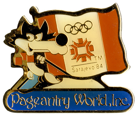 Enameled button in the shape of Vuchko, the 1984 Sarajevo Winter Olympics wolf mascot, on skis next to the flag depicting the logo for the games. Text reads "Sarajevo '84 Pageantry World, Inc."