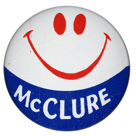 Round, white button with a curving band of blue along the bottom and a red smiley face in the upper half. White text reads "McCLURE"