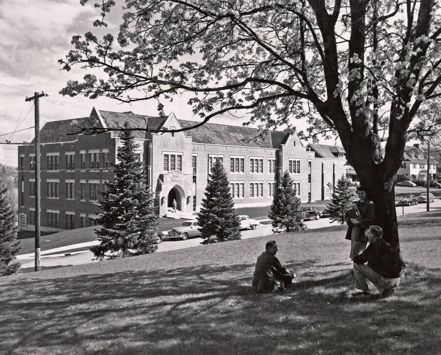 1951 photograph of the Music Building. Three students sit in the shade in the foreground. [PG1_117-01]