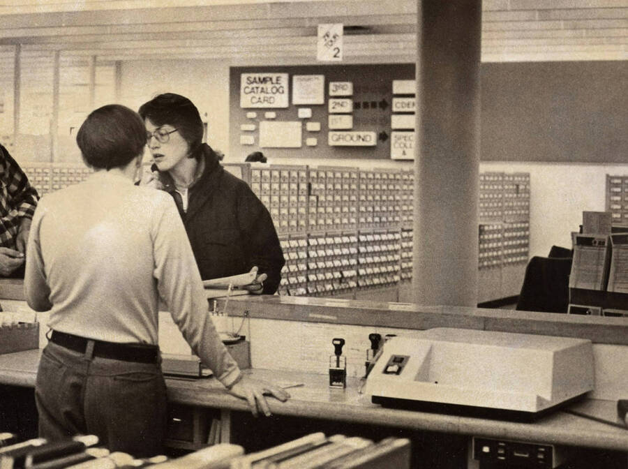 1978 photograph of the Library. A library employee helps a student at the loan desk with card catalog in the background. [PG1_122-082a]