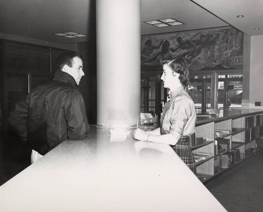 1957 photograph of the Library. A library employee talks to a student at the loan desk. [PG1_122-017]