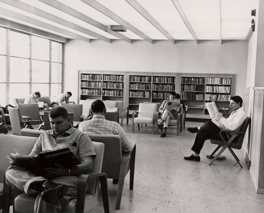 1957 photograph of the Library. Students browse newspapers in foreground, bookshelf in background. [PG1_122-020]