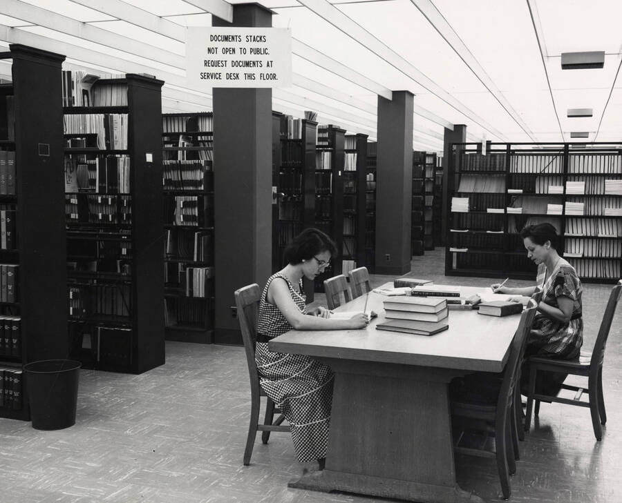 1957 photograph of the Library. Government Documents. Two students work at a table in foreground. [PG1_122-048]