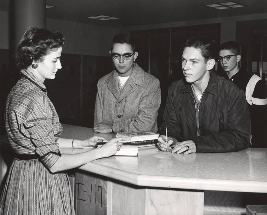 1957 photograph of the Library. Library employee assist students at loan desk. Donor: Publications Dept. [PG1_122-073]