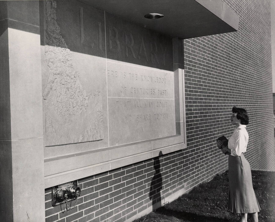 1957 photograph of the Library. A student looks at the entrance plaque to the library. 'Here is the knowledge of centuries past that all may come and share today.' Donor: Publications Dept. [PG1_122-080]