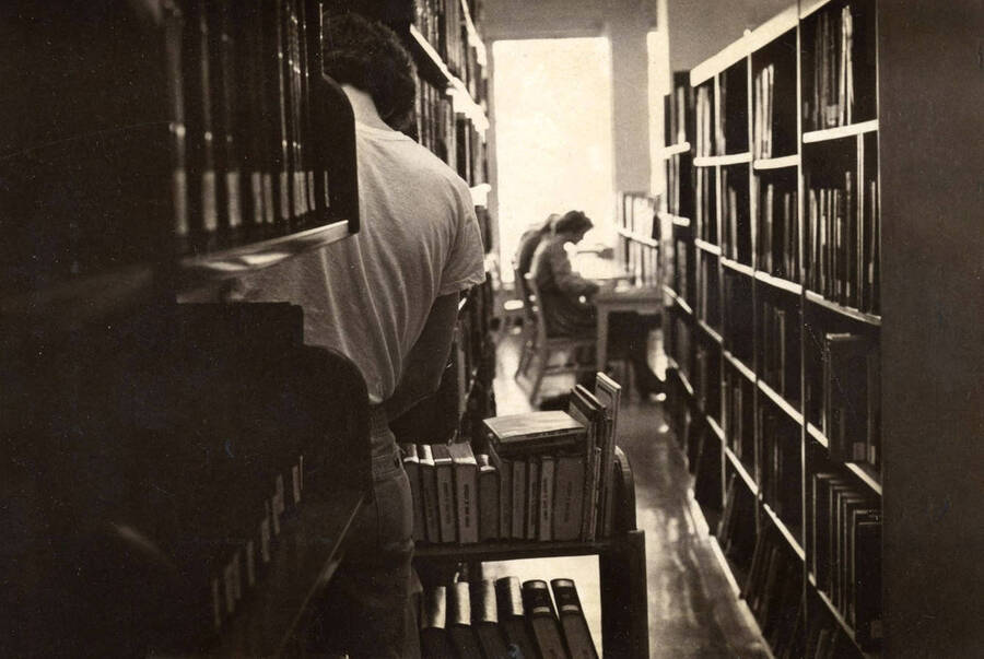 1978 photograph of the Library. A library employee shelves books while students study in the background. [PG1_122-087]