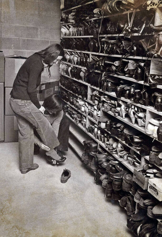 1974 photograph of Hartung Theatre. Two students try on shoes in the costume storage area. [PG1_145-07]