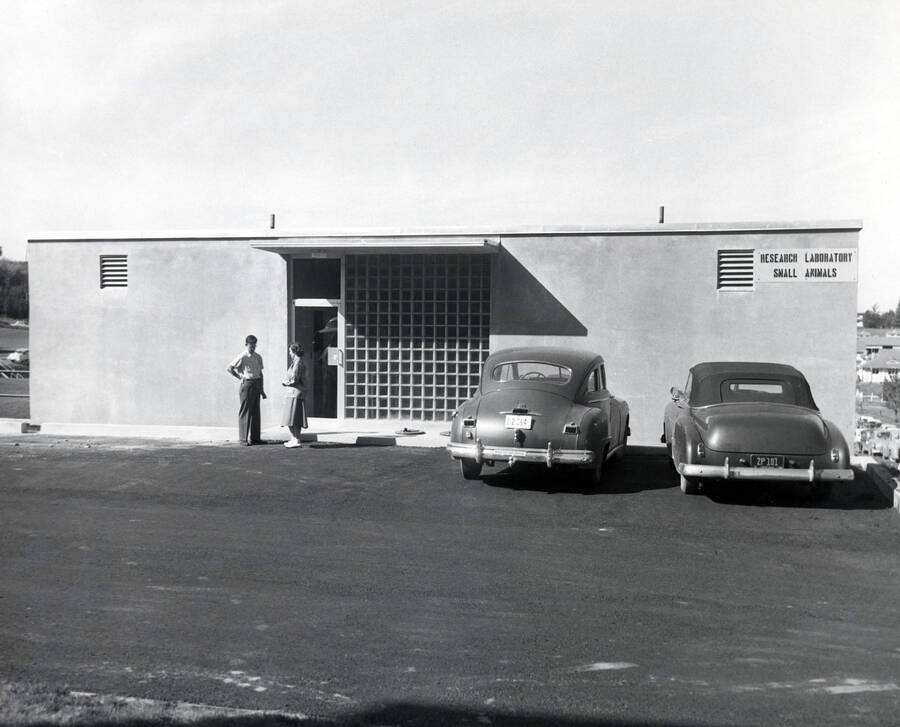 1946 photograph of the Small Animal Research Laboratory. Two students and automobiles in foreground. Donor: Publications Dept. [PG1_156-02]