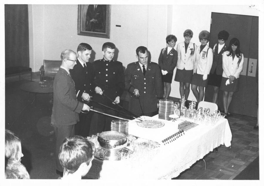 1969 photograph of Military Science Cadets. Four cadets cut into a cake with swords at a Tri-Service ceremony. Donor: Publications Dept. [PG1_208-122]