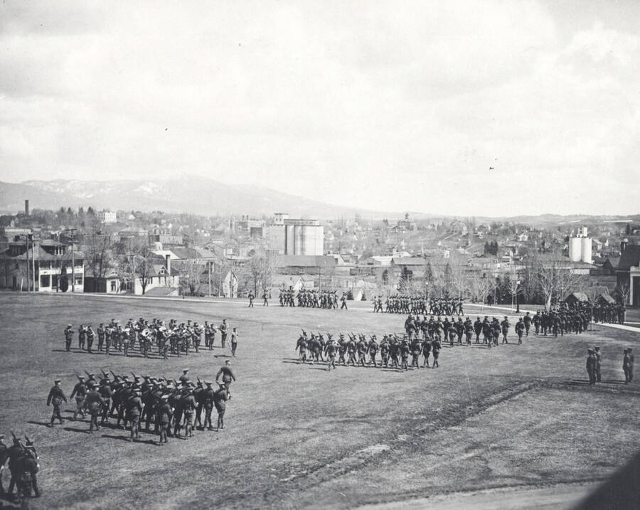 1914 photograph of Military Science Cadets. Military cadet regiments in review with campus buildings in the background. [PG1_208-022]