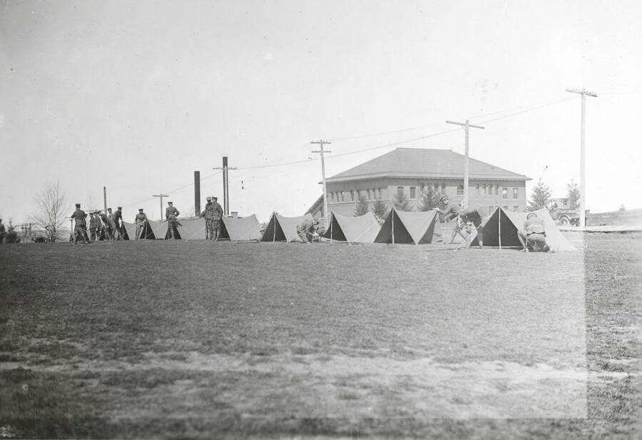 1922 photograph of Military Science Cadets. Military cadets setting up tents on campus. Campus buildings in the background. [PG1_208-024]