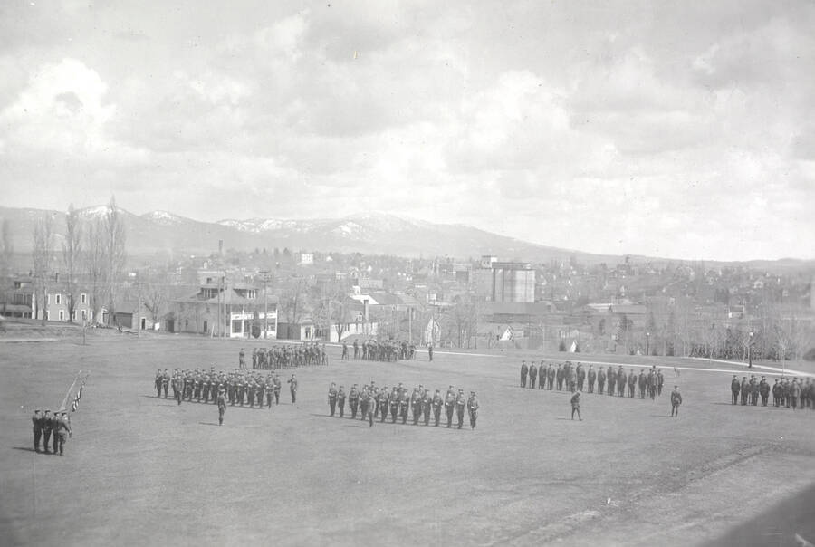 1922 photograph of Military Science Cadets. Military cadets on parade on campus grounds. Campus buildings in the background. [PG1_208-027]