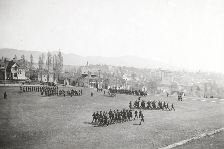 1922 photograph of Military Science Cadets. Military cadets on parade on campus grounds. Campus buildings in the background. [PG1_208-028]