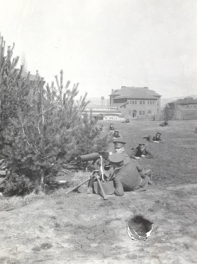 1914 photograph of Military Science Cadets. Military cadets participate in a machine gun drill. Campus buildings are visible in the background. [PG1_208-046]