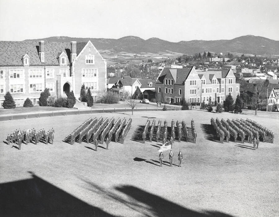 1945 photograph of Military Science Cadets. Military cadets in parade formation on campus grounds with buildings in the background. [PG1_208-054]
