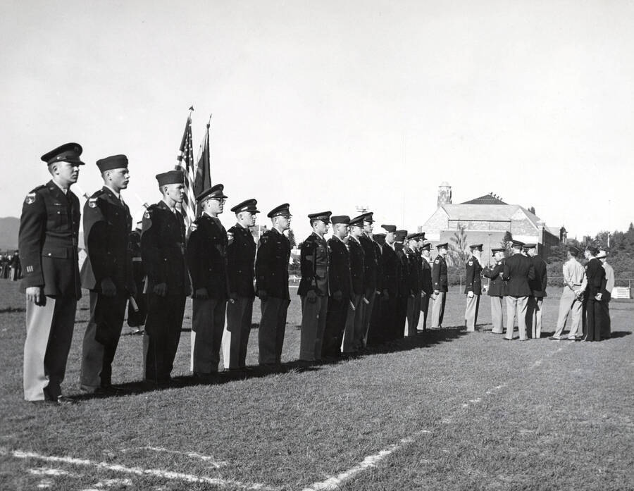 1953 photograph of Military Science Cadets. Cadet receiving an adward at MacLean Field. Campus buildings visible in the background. [PG1_208-079]