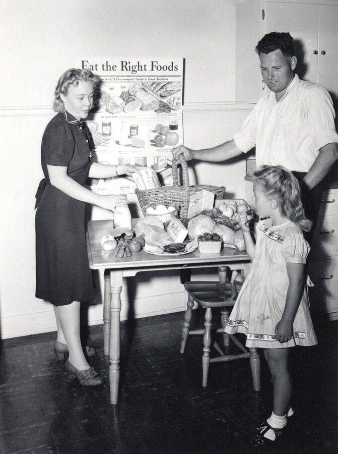 1953 photograph of Home Economics. A table with food in front of a poster that reads 'Eat the Right Foods.' [PG1_221-109]