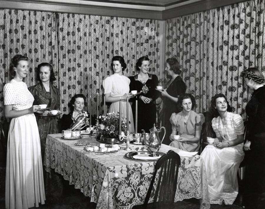 1945 photograph of Home Economics. Students engage in a formal tea around a set table. [PG1_221-018]