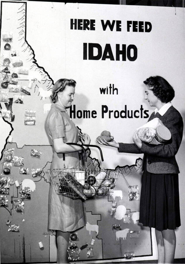 1941 photograph of Home Economics. Two students with Idaho grown products stand in front of a large poster of Idaho with the legend 'Here we feed Idaho with home products.' Donor: Publications Dept. [PG1_221-062]