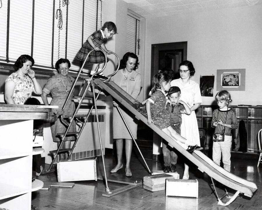 1965 photograph of Home Economics. Students watch children playing on a slide. Donor: Publications Dept. [PG1_221-078]
