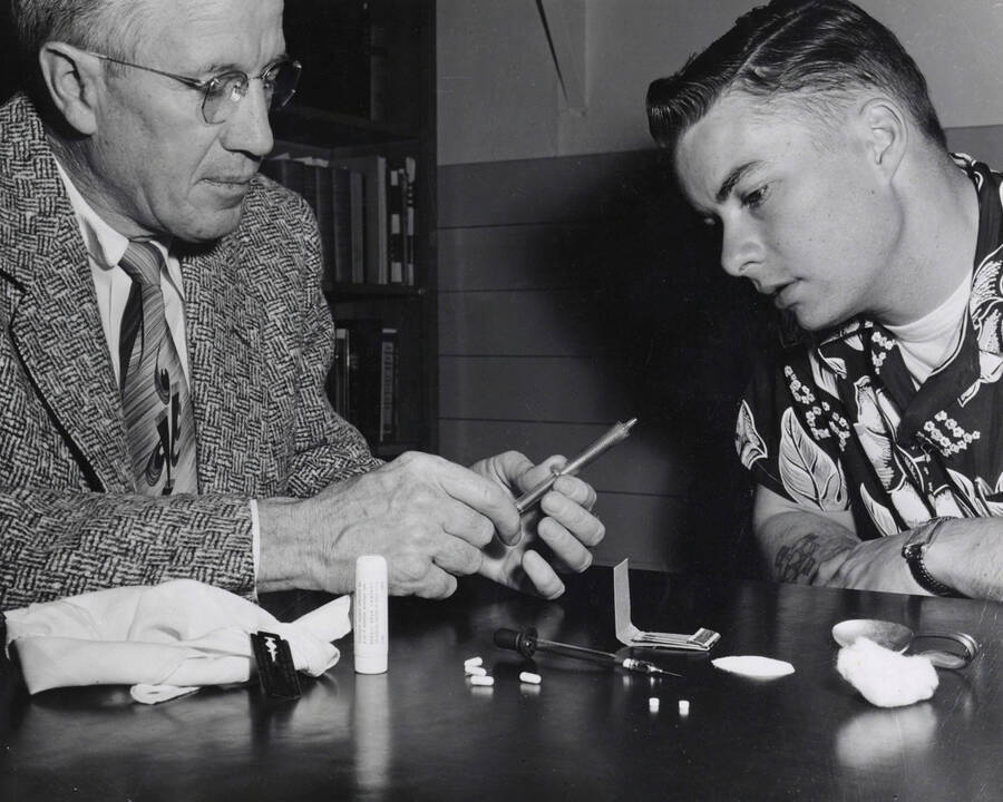 1954 photograph of Sociology Buckled. Prof. Harry C. Harmsworth and Edwin Henry Pfuhl Jr. examine drug paraphernalia [PG1_244-01b]