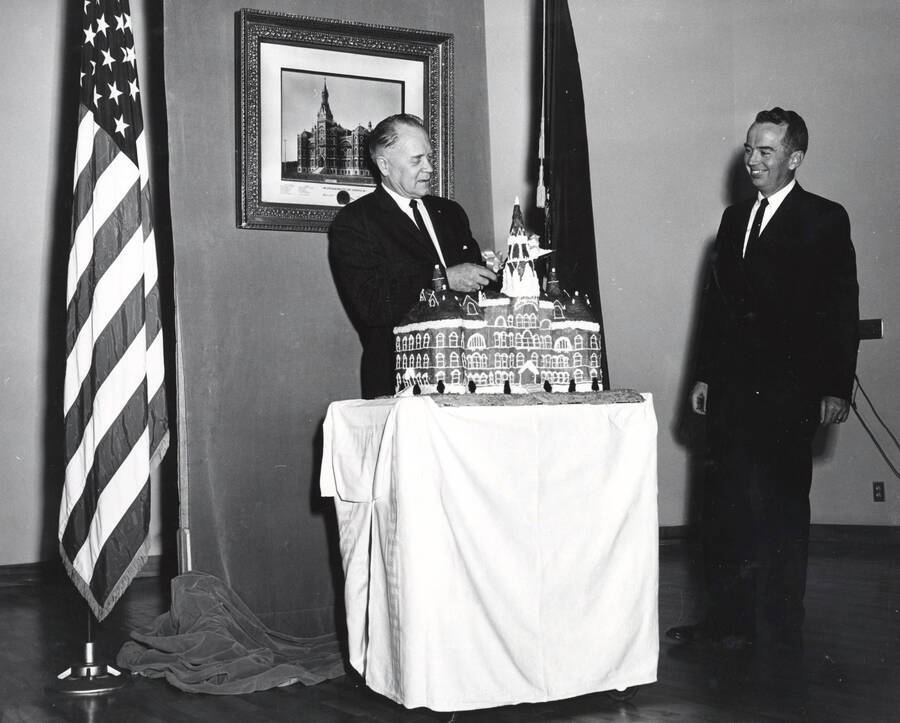 1964 photograph of 75th Anniversary. Ezra M. Hawkes and James H. Roper cuttingt a cake shaped like the old Administration building. [PG1_246-18b]