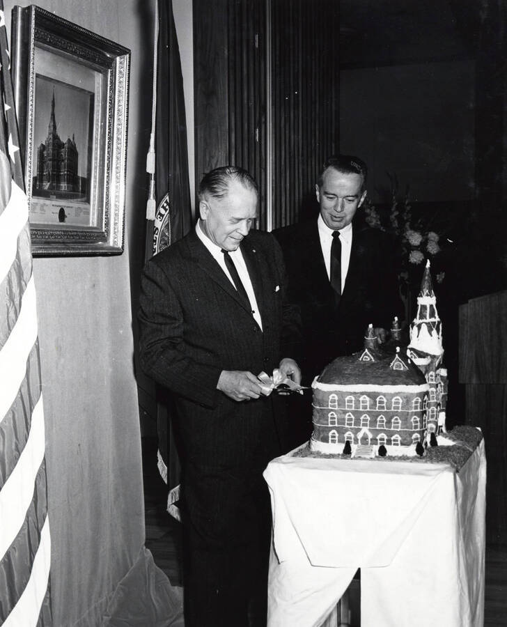 1964 photograph of 75th Anniversary. Ezra M. Hawkes and James H. Roper cuttingt a cake shaped like the old Administration building. [PG1_246-18c]