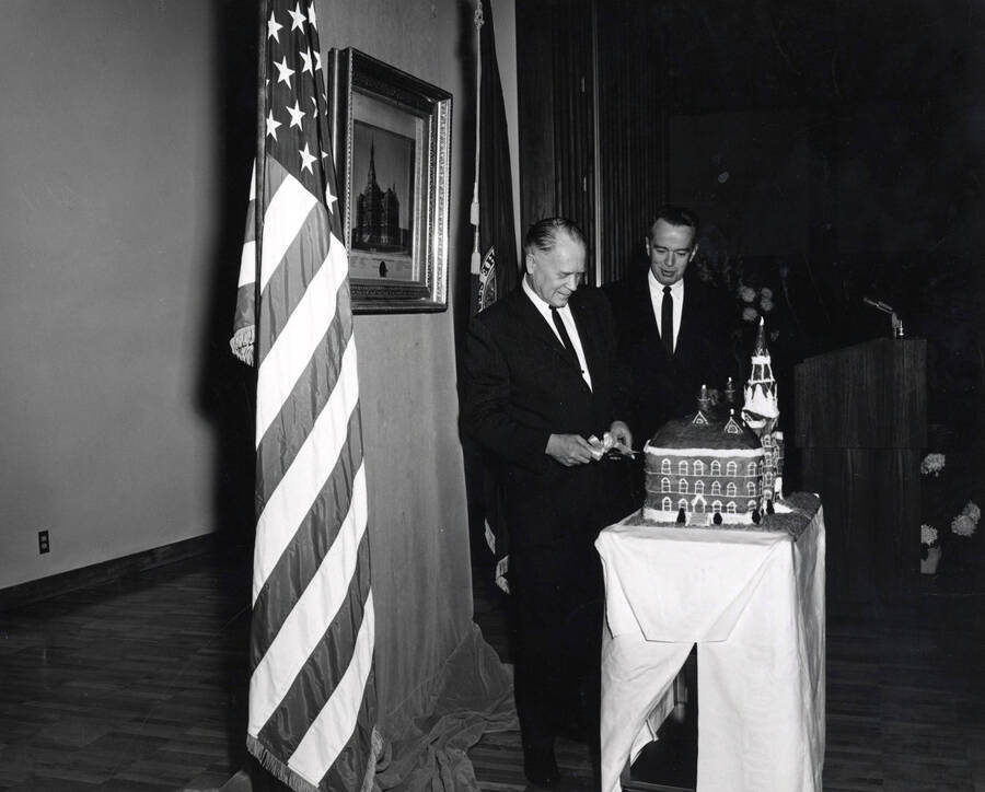 1964 photograph of 75th Anniversary. Ezra M. Hawkes and James H. Roper cuttingt a cake shaped like the old Administration building. [PG1_246-18e]