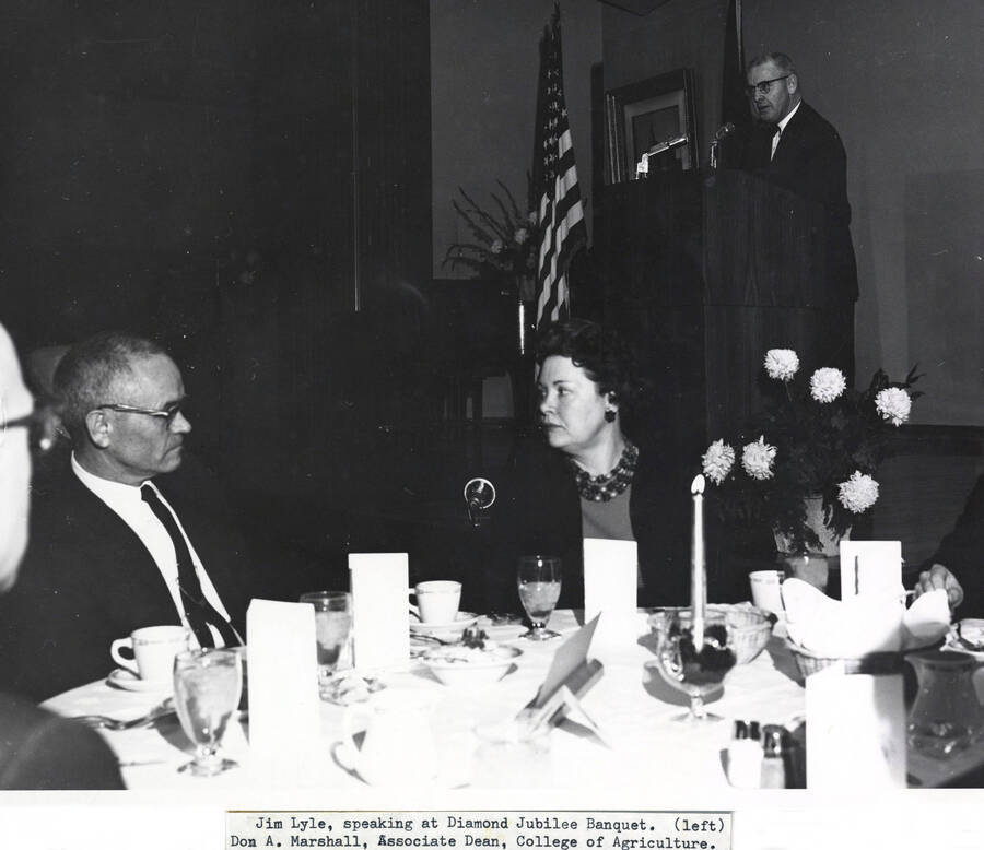 1964 photograph of 75th Anniversary. Don Marshall seated at a table with Jim Lyle standing at a lecturn in the background during the Diamond Jubilee. [PG1_246-19]