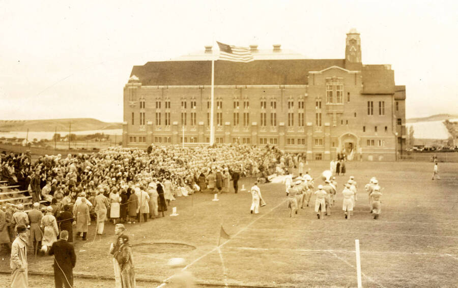 1930 photograph of Memorial Gymnasium. Marching band shown on the field with the fans in the grandstands. [PG1_61-05]