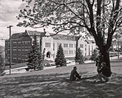 1951 photograph of the Music Building. Three students sit in the shade in the foreground.