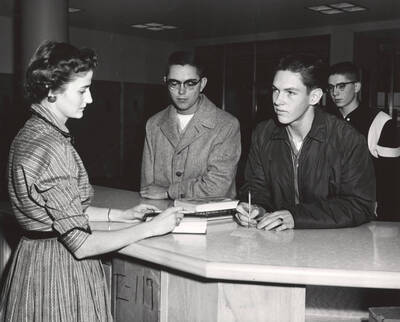 1957 photograph of the Library. Library employee assist students at loan desk. Donor: Publications Dept.