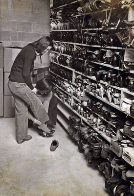 1974 photograph of Hartung Theatre. Two students try on shoes in the costume storage area.