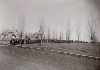 1897 panoramic photograph of University of Idaho campus. Students lined up for procession on sidewalk. Donor: Homer David.
