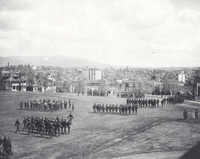 1914 photograph of Military Science Cadets. Military cadet regiments in review with campus buildings in the background.