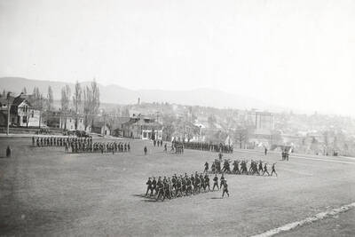 1922 photograph of Military Science Cadets. Military cadets on parade on campus grounds. Campus buildings in the background.