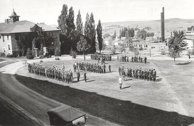 1925 photograph of Military Science Cadets. New cadet recruits at inspection in front of gymnasium with campus buildings in the background.