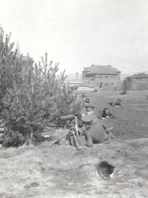 1914 photograph of Military Science Cadets. Military cadets participate in a machine gun drill. Campus buildings are visible in the background.